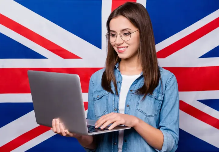 Uśmiechnięta dziewczyna w okularach pracuje na laptopie na tle flagi Wielkiej Brytanii, tworząc przykładowy artykuł po angielsku.
