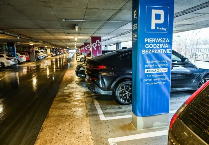 Mokry parking nad zaporą, rząd samochodów pod betonowym stropem. Niebieska tablica informuje o pierwszej darmowej godzinie.