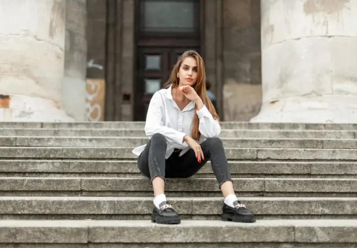 Eleganckie mokasyny to idealne buty do szarych spodni. Kobieta w białej koszuli i czarnych jeansach siedzi na schodach.