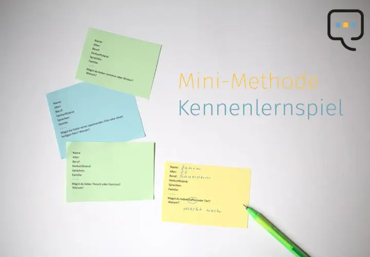 Bunte Zettel mit Fragen für ein Kennenlernspiel liegen auf weißem Grund. Ein grüner Stift liegt daneben.