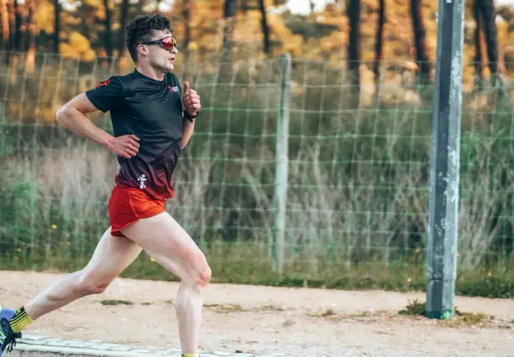 Mężczyzna w sportowym stroju biegnie, ćwicząc jak zwiekszyc kadencje w bieganiu. W tle siatka i drzewa.
