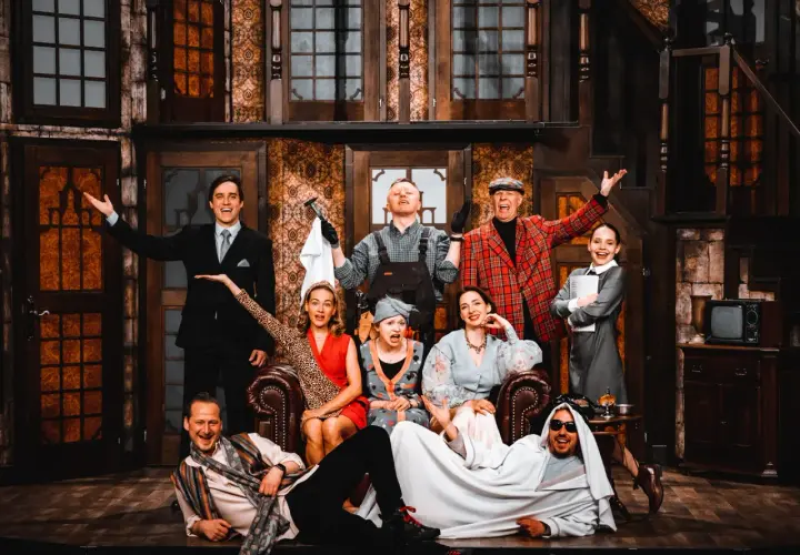 Aktorzy w teatralnej scenografii, jakby w teatrze bez rzędów, pozują do zdjęcia. Wszyscy uśmiechnięci, w barwnych strojach, tworzą radosną atmosferę.