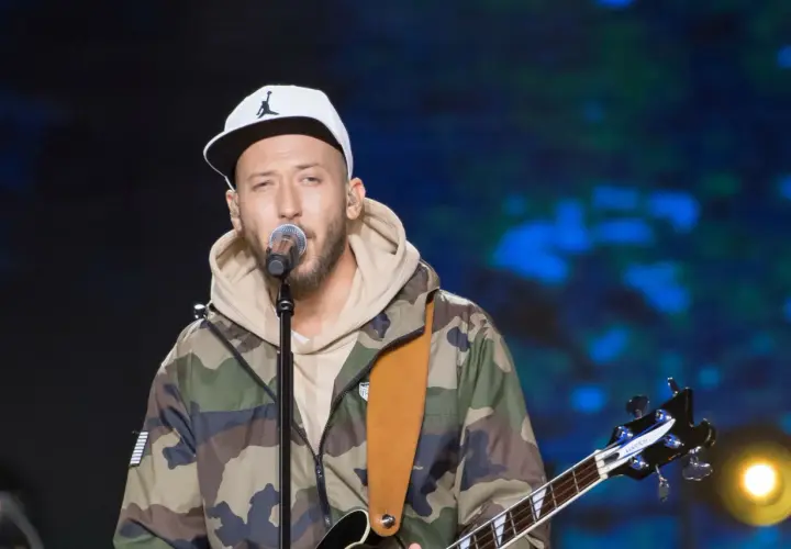 Artysta w czapce z daszkiem i kurtce moro śpiewa do mikrofonu, trzymając gitarę. Może to jego żona czeka na niego po koncercie?
