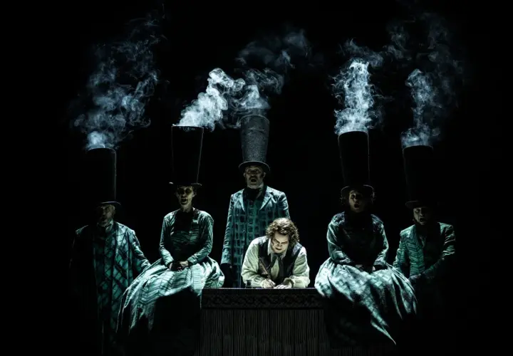 Scen från Sweeney Todd operan. Människor med höga hattar och rök som stiger ur dem. En man skriver vid ett bord.
