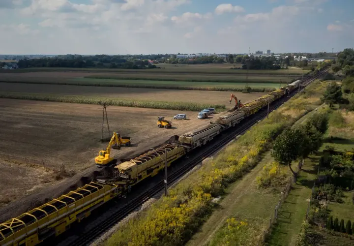 Długi pociąg z wagonami wypełnionymi ziemią, obok koparki i samochodów. Prace torowe w tle pól i zabudowań, widać odbojnica kolejowa.