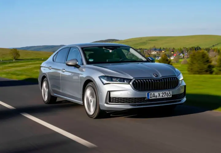 Srebrna, nowa Skoda Superb pędzi po drodze, mijając malownicze, zielone wzgórza i domki.