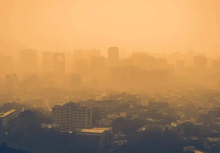 Vista aérea de una ciudad envuelta en una densa neblina anaranjada, con siluetas de edificios apenas visibles. Las partículas PM10 flotan en el aire, creando esta atmósfera opresiva.