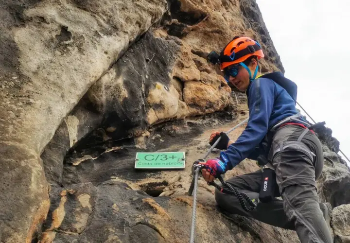 Młoda osoba w kasku wspina się po skale na via ferrata w Decinie. Tabliczka "Cesta do nebíčka" wskazuje trudność trasy.