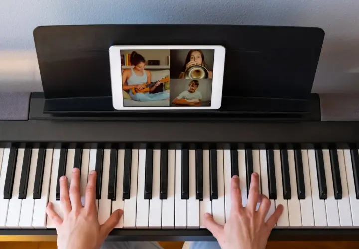 Ręce na klawiaturze pianina cyfrowego, na ekranie tabletu widać lekcję muzyki online z uczniami grającymi na ukulele, trąbce i gitarze.