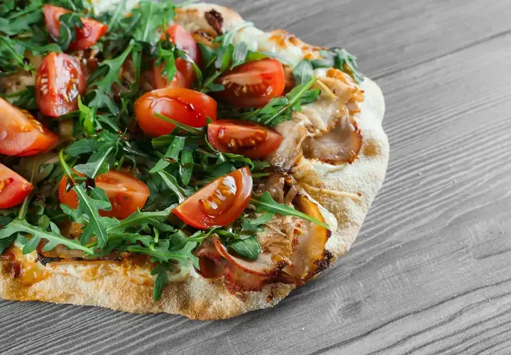 Une délicieuse pinsa recette garnie de tomates cerises, roquette fraîche et viande effilochée, sur fond de bois gris.