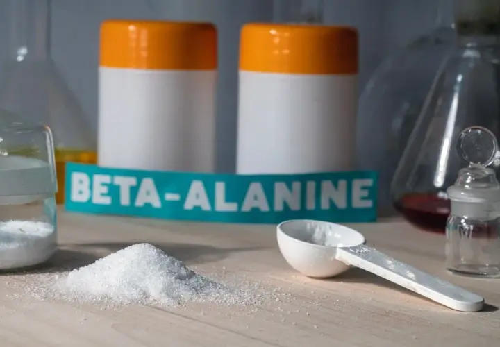 Biały proszek beta-alaniny na blacie, obok miarki i słoiczka. W tle dwa białe pojemniki z pomarańczowymi nakrętkami. Czy beta-alanina skutki uboczne są powodem do obaw?