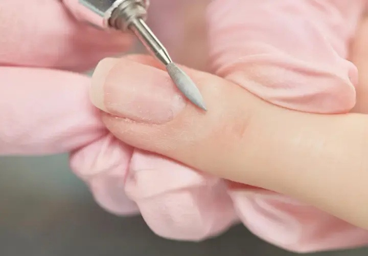 Dłoń w różowej rękawiczce usuwa frezarką resztki lakieru hybrydowego z paznokcia.