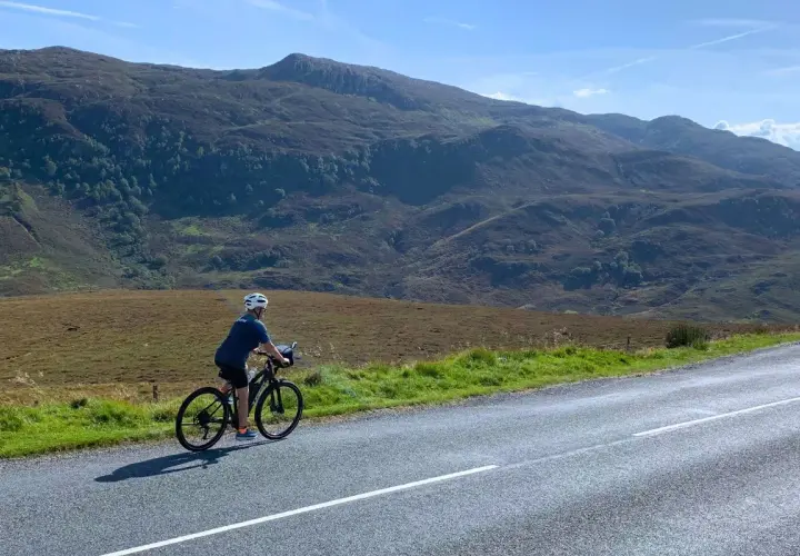 A lone cyclist enjoys a scenic bike tour Scotland, with rolling hills and mountains in the background under a bright blue sky.