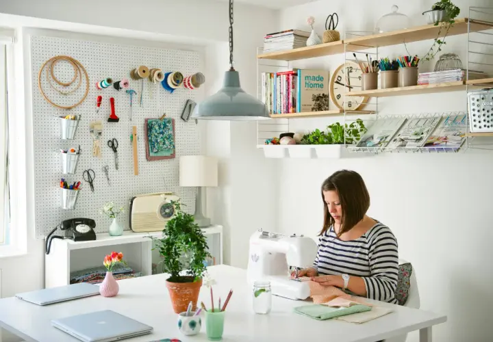 Mujer cosiendo en su **taller de manualidades en casa**. Pared con herramientas, estanterías con libros y plantas.