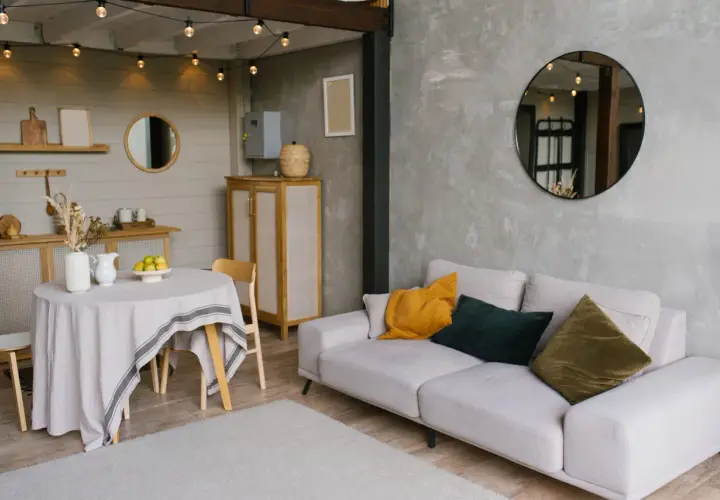 Un salon moderne avec un canapé clair, des coussins colorés et un miroir rond. La table ronde couverte d'une nappe grise et les guirlandes lumineuses ajoutent une touche chaleureuse, reflétant une nouvelle tendance déco.