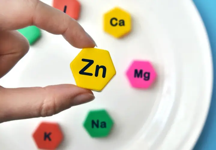 Palec wskazujący trzyma żółty heksagon z napisem "Zn". Wokół niego na białym talerzu leżą inne heksagony z symbolami pierwiastków: Ca, Mg, Na, K.
