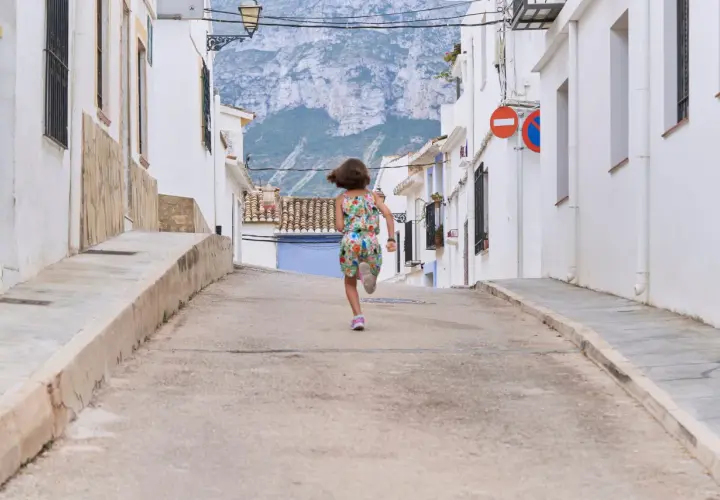 Niña corre por calle empinada de un pueblo de Alicante, con casas blancas y montañas al fondo.