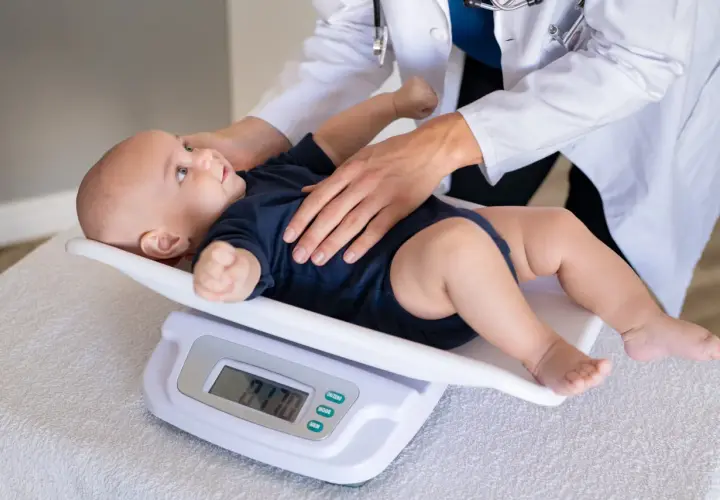 Bebé de 11 meses en báscula digital, con manos de doctor. El peso del bebé es de 9.70 kg.