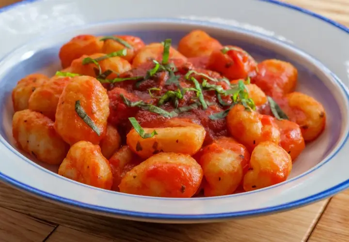Pyszne gnocchi z sosem pomidorowym i świeżą bazylią, podane w białej misce z niebieskim rantem.