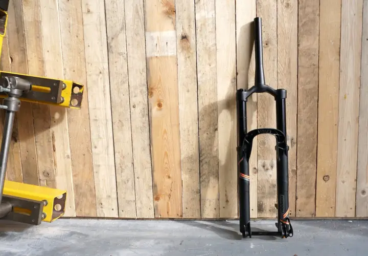 A black MRP Ribbon coil suspension fork stands against a wooden wall. The fork features orange and grey accents and the "ribbon" branding is visible.