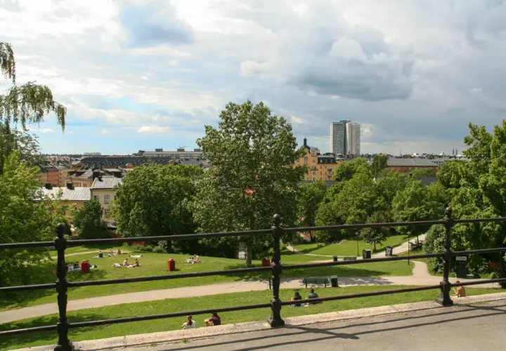 Sommar i Vitabergsparken, Stockholm. Människor njuter av solen på gräsmattan, med stadens takåsar och en teater i bakgrunden.