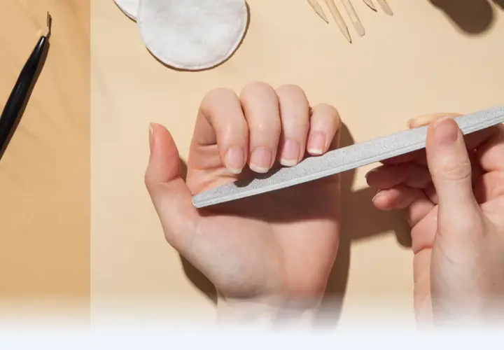 Dłonie pielęgnują paznokcie, używając pilnika. W tle widać narzędzia do manicure, takie jak nożyczki i cążki, tworząc kompletny zestaw do pielęgnacji macierzy paznokcia.