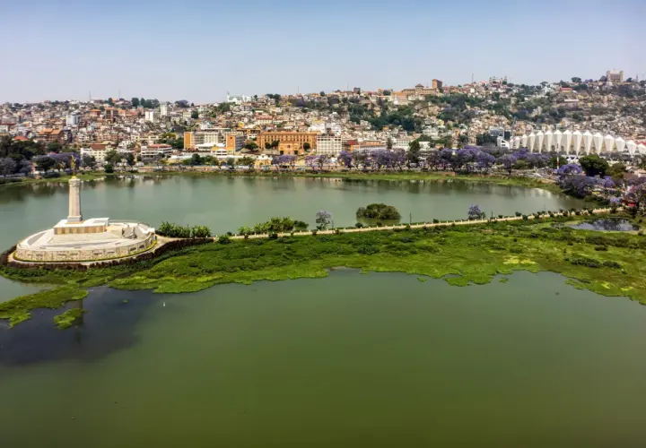 Widok na jezioro i wzgórza Antananarywy, gdzie żyje madagaskar ludność. Widać pomnik, stadion i kwitnące drzewa.