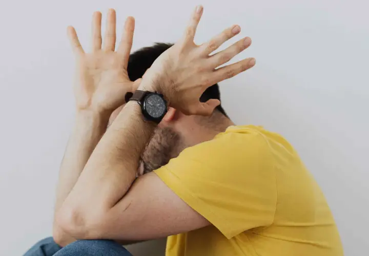 Uomo con le mani alzate a protezione del viso, forse per la paura delle cimici. Indossa una maglietta gialla e un orologio.