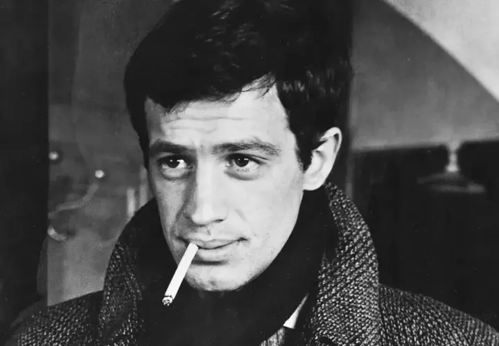 Jean-Paul Belmondo z papierosem w ustach, w płaszczu. Jego spojrzenie przywodzi na myśl niezapomniane **belmondo cytaty**.