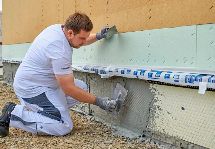Handwerker kniet und verputzt die Sockelzone einer Hausfassade mit einer Kelle.