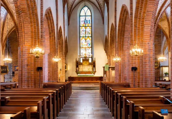 Interiör av Helga Trefaldighets kyrka med bänkar, tegelvalv och ett färgstarkt glasmålat fönster bakom altaret.
