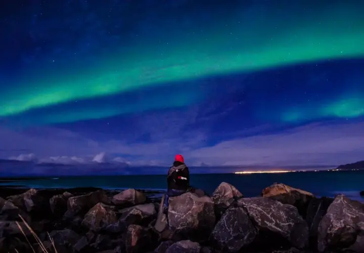 Person sitzt auf Felsen am Meer und blickt auf Nordlichter. Perfektes Wetter auf Island im September für dieses Naturschauspiel.
