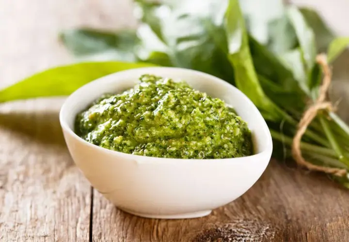 Pesto verde in una ciotola bianca, con un fascio di foglie di sedano fresche sullo sfondo. Perfetto per le tue ricette.