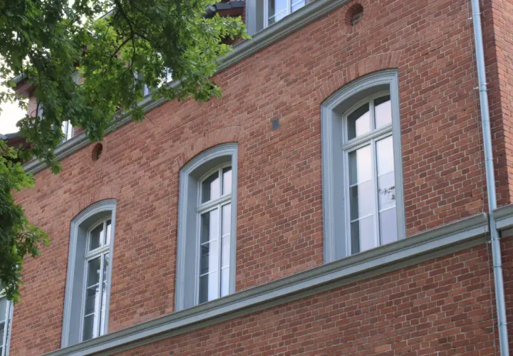 Eleganckie okna drewniane zabytkowe w ceglanej kamienicy, otoczone zielenią drzew.