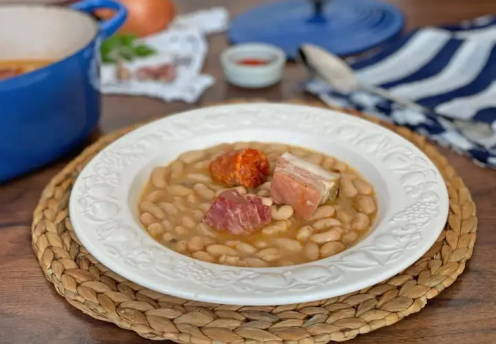Plato de judías blancas con chorizo y tocino, servido en una vajilla blanca sobre una esterilla de mimbre.