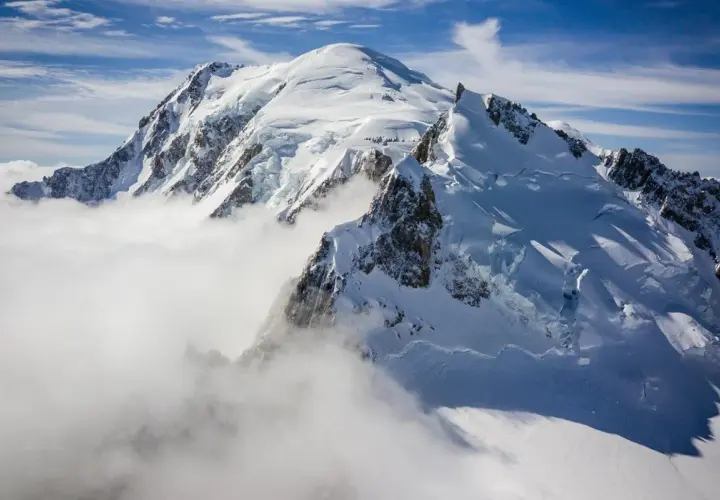 Le Mont Blanc, majestueux et enneigé, émerge des nuages. Des alpinistes escaladent ses pentes glacées sous un ciel bleu éclatant.