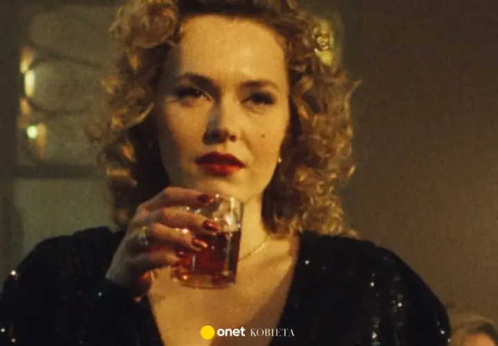Kobieta z burzą blond loków i czerwoną szminką, pijąca drinka. W tle widać fragmenty scenografii, sugerujące klimat filmu "Jak pokochałam gangstera obsada".