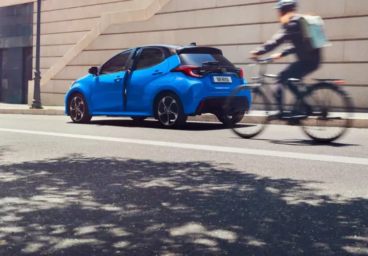 Una Toyota Yaris blu brillante sfreccia in città, mentre un ciclista con zaino passa velocemente. Diversi modelli di Toyota Yaris sono disponibili per ogni stile di vita.