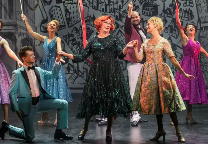 Scen från "Tootsie" på Oscarsteatern. En man i ljusblå kostym knäböjer medan en kvinna i glittrig grön klänning och andra i färgglada klänningar dansar.