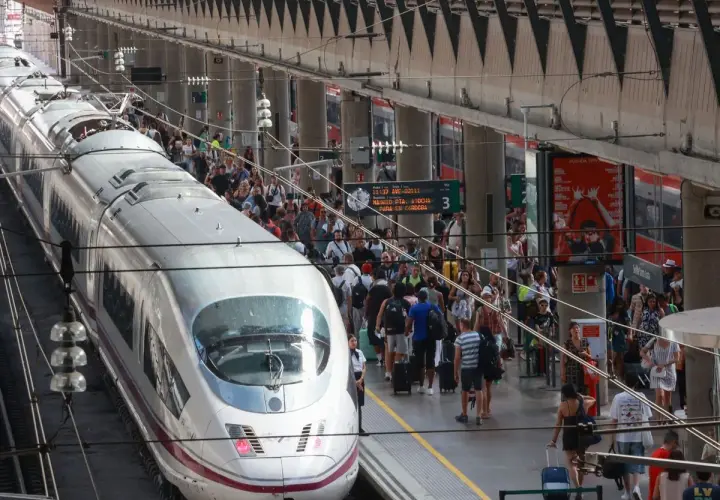Un tren AVE espera en la estación, listo para llevar pasajeros de Lleida a Sevilla. Gente con maletas se agolpa en el andén.