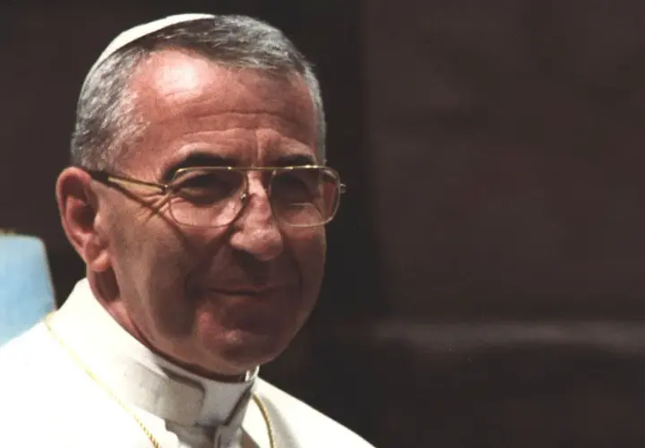 Porträt von Papst Johannes Paul I. mit Brille und Soutane, im Hintergrund eine goldene Statue.