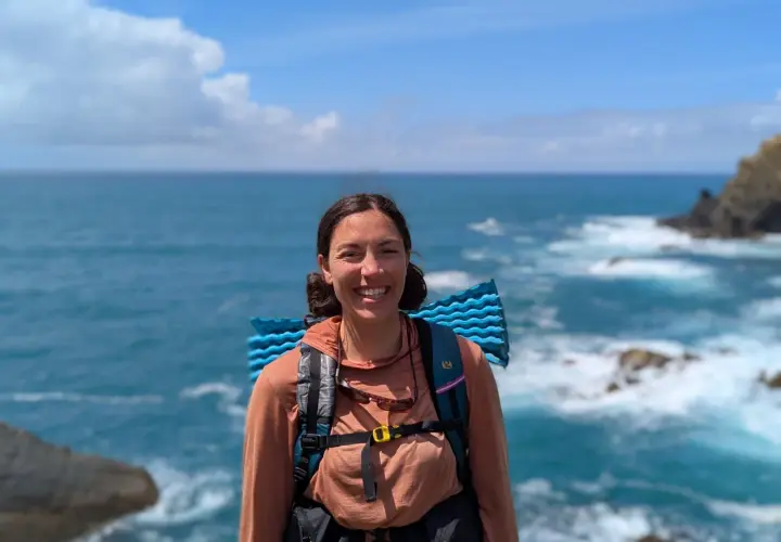 A smiling hiker with a backpack and sleeping pad enjoys the view of the ocean waves crashing on the shore along the Fischerweg trail in Portugal.