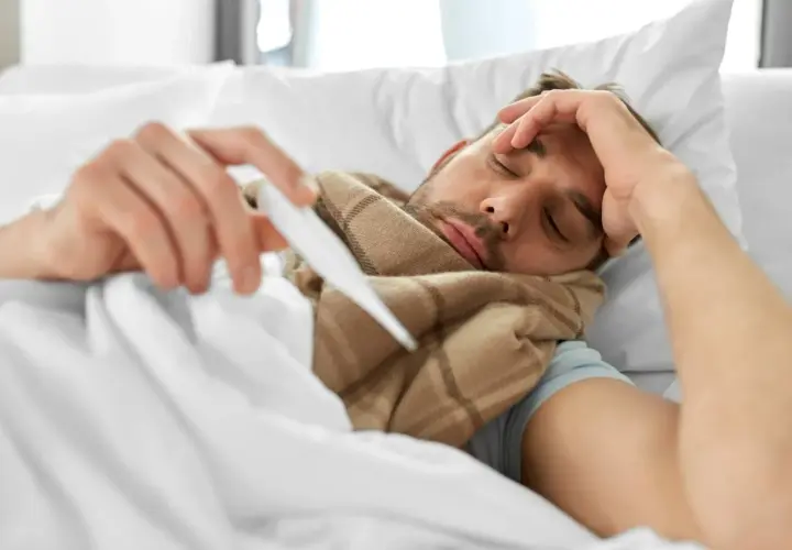 Chory mężczyzna leży w łóżku, trzymając termometr. Zastanawia się, co na gorączkę.