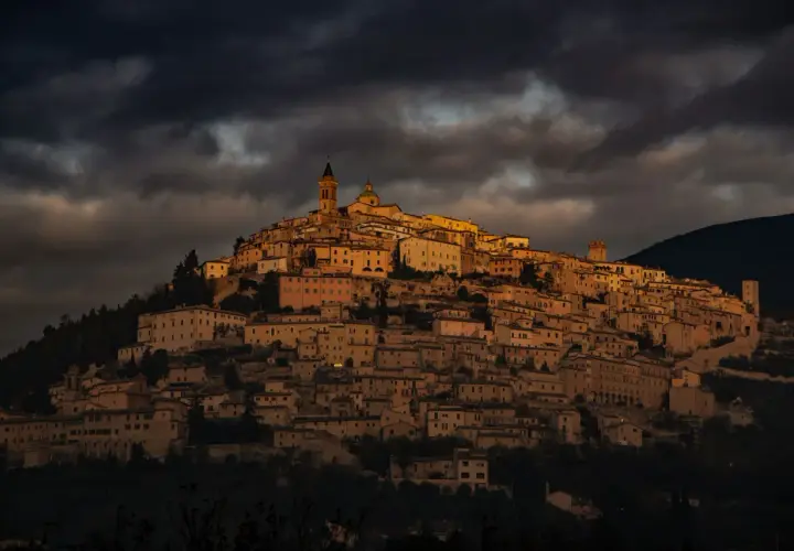 Antico borgo di Trevi, Umbria, illuminato da un sole dorato che filtra tra nuvole scure. Case in pietra si aggrappano alla collina.
