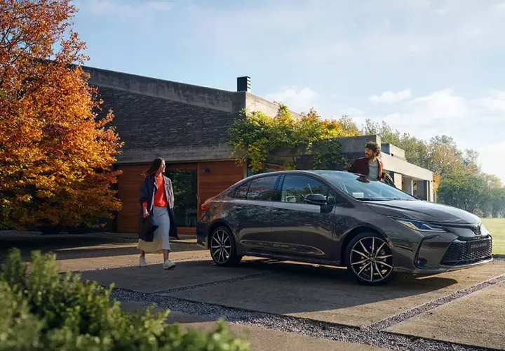 Un hombre y una mujer junto a un Toyota Corolla oscuro frente a una casa moderna.
