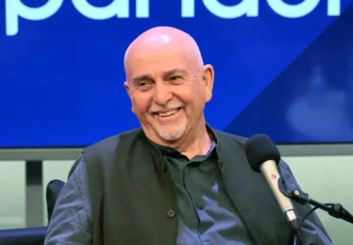 Uśmiechnięty Peter Gabriel, legendarny wokalista, w studiu. W tle niebieskie logo, przypominające o jego bogatej karierze z zespołem Genesis.
