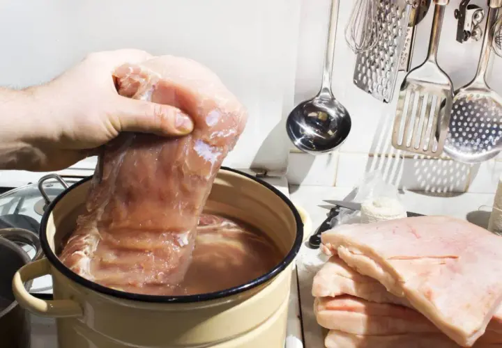 Przygotowanie solanki do mięsa: ile soli na litr wody? Duży kawałek mięsa zanurzany w wodzie w garnku, obok stos słoniny i kuchenne akcesoria.
