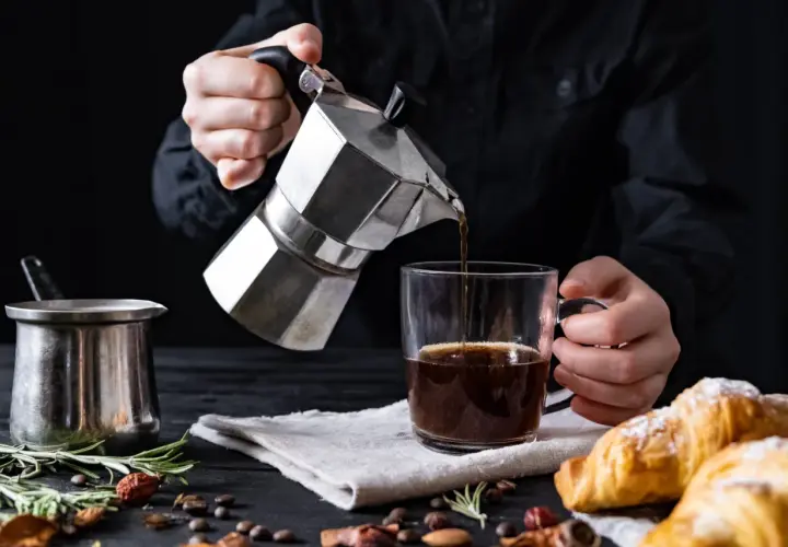 Cafetière italienne inox versant du café dans une tasse. Un petit pot en métal et des croissants sont sur la table.
