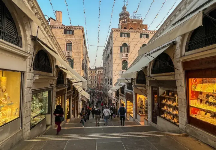 Schody na moście Rialto w Wenecji, z witrynami sklepów z biżuterią po bokach. Tłum ludzi schodzi w dół.
