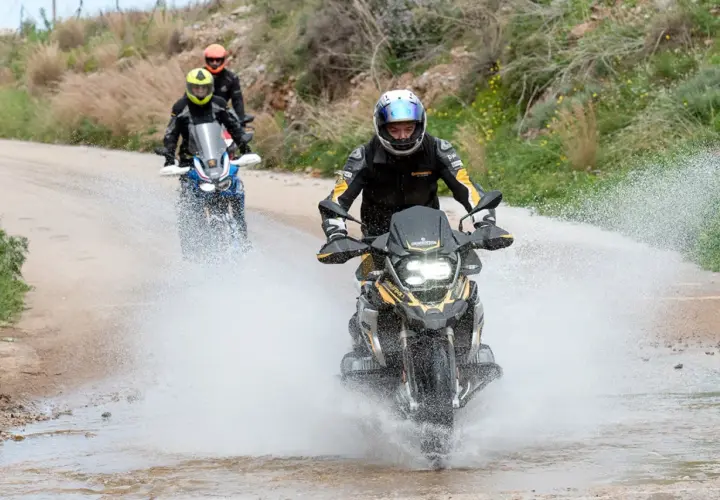 Motociclistas en un **conti trail attack 4 test** atraviesan un charco de agua en un camino de tierra, levantando una gran estela.